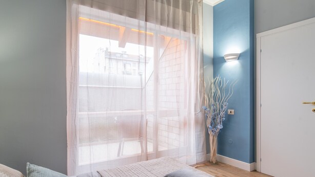 Un dormitorio con cama, una ventana con cortinas translúcidas, un jarrón con ramas y una lámpara fijada a la pared.