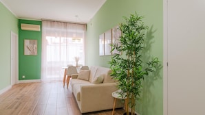 Una sala de estar con un sofá color beige, una mesa de comedor con sillas, una planta en maceta y un ventanal con cortinas translúcidas.