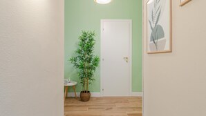 Un pasillo con pared verde, una puerta blanca, una planta de bambú en maceta, una mesita redonda con un objeto blanco, y una obra de arte enmarcada.