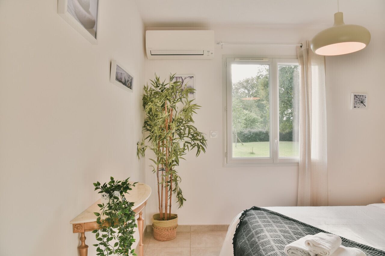 A bedroom with a bed, a window with curtains, a wall-mounted air conditioner, and a potted plant.