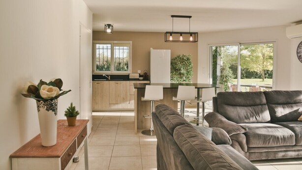 A modern living room with a grey sofa, a wooden side table with a vase, and a view of the kitchen and garden through large windows. - House for 6, swimming pool and garden (Bénesse-Maremne)