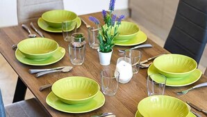 A dining table set with green plates, silverware, and clear glasses, with a small potted plant in the center. -  Home Sweet Home (Monreale)
