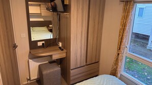 A compact bedroom with a wooden wardrobe, a bedside table with a remote control, a small stool, and a window with curtains. - 3br-sleeps6-parking-pool-nearbeach&skegness (Skegness)
