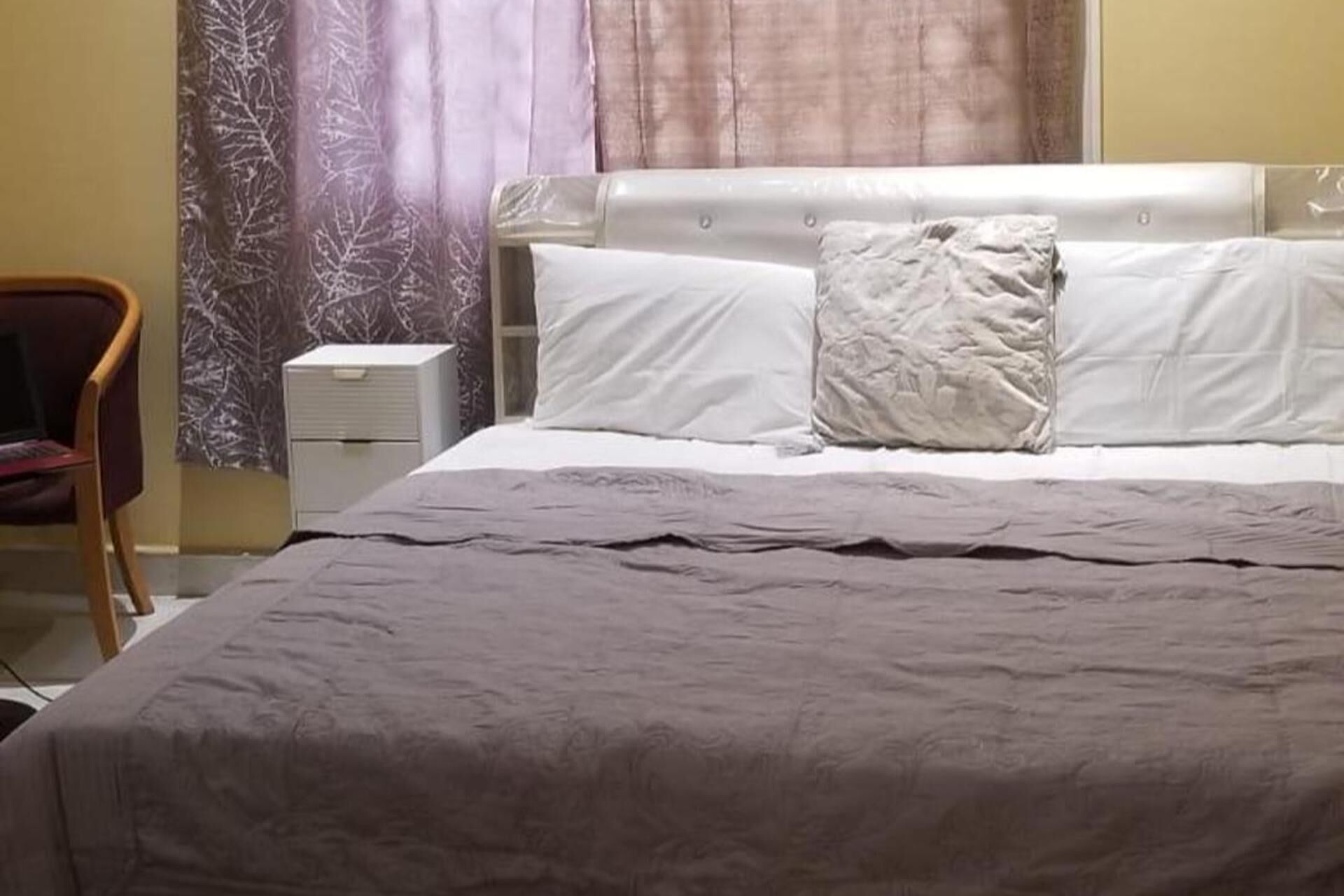 Ein ordentlich bezogenes Bett mit grauer Tagesdecke und weißen Kissen, ein Holzsessel und ein kleiner Nachttisch mit Lampe.
