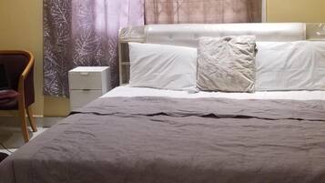 Ein ordentlich bezogenes Bett mit grauer Tagesdecke und weißen Kissen, ein Holzsessel und ein kleiner Nachttisch mit Lampe.