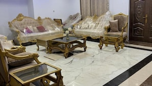 Una habitación con un sofá grande y ornamentado, una mesita de centro y una silla. El suelo está cubierto de baldosas y la puerta presenta elaborados detalles de madera.