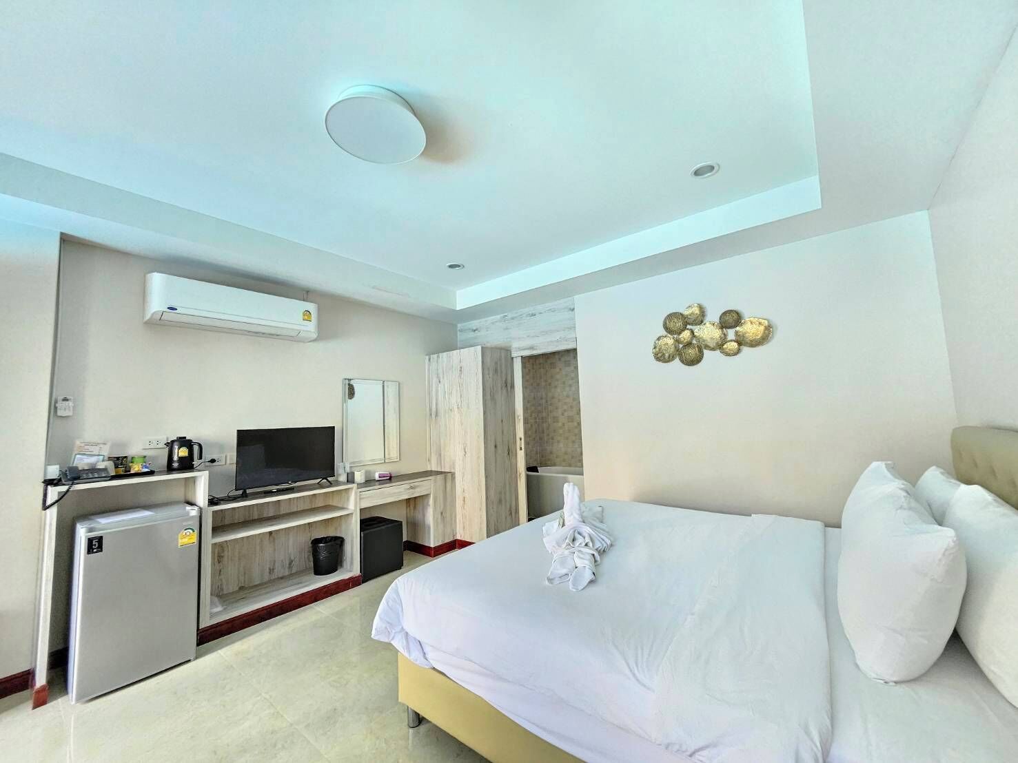 A hotel room with a bed, a desk with a television, a mini-fridge, and an air conditioning unit.