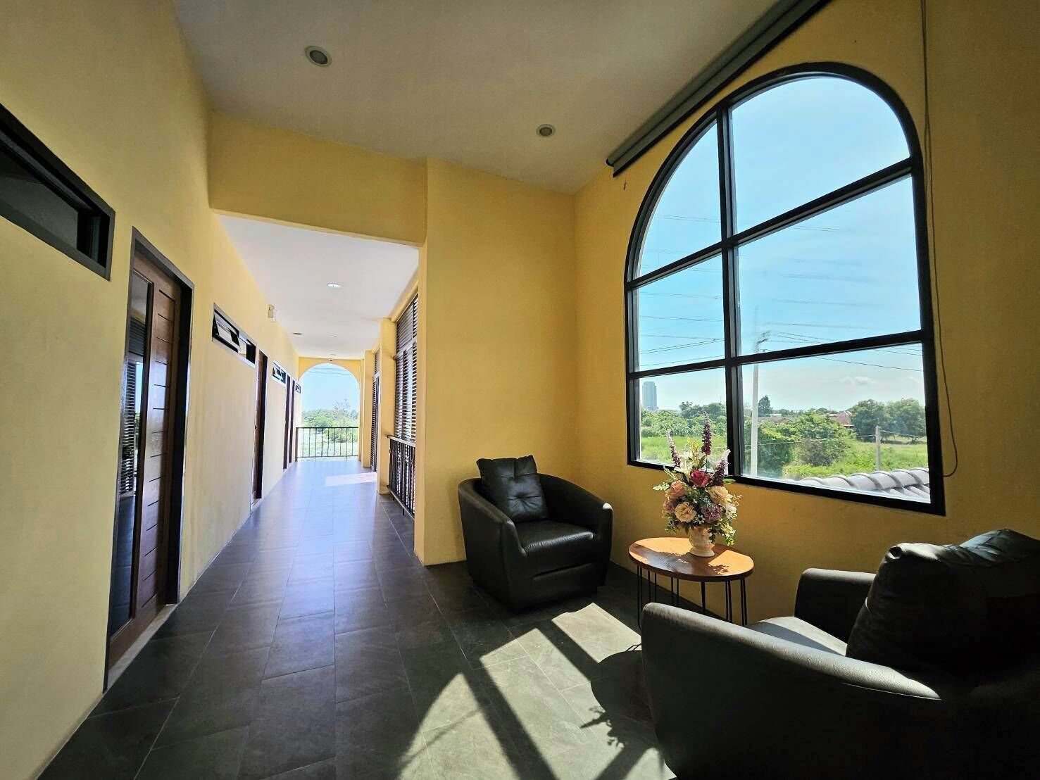 A hotel corridor with a long view to the outside, a black leather sofa, a small round table with a vase, and a large arched window.