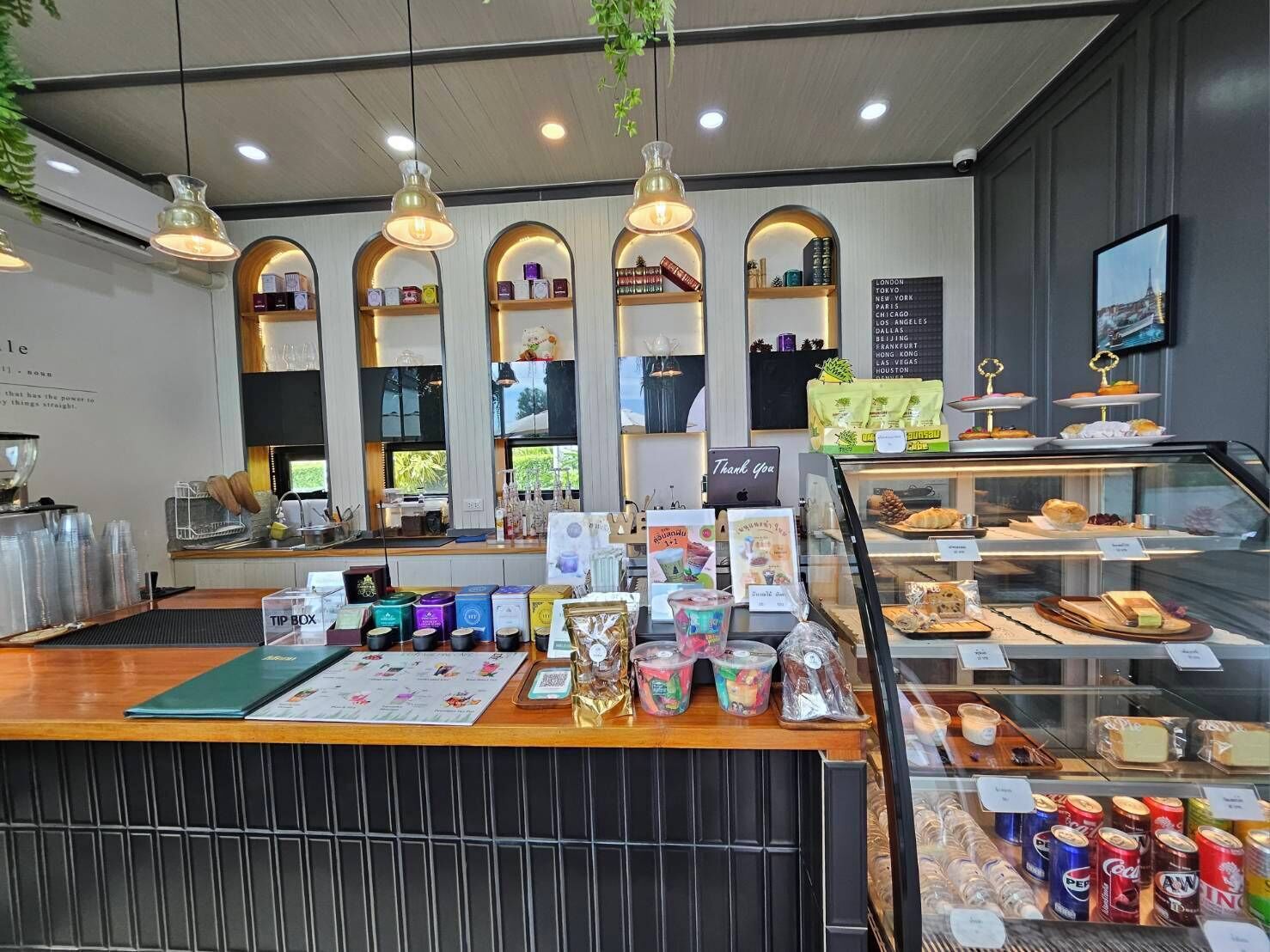 Una cafetería con mostrador de madera, una vitrina con pasteles y una pizarra que enumera ciudades.