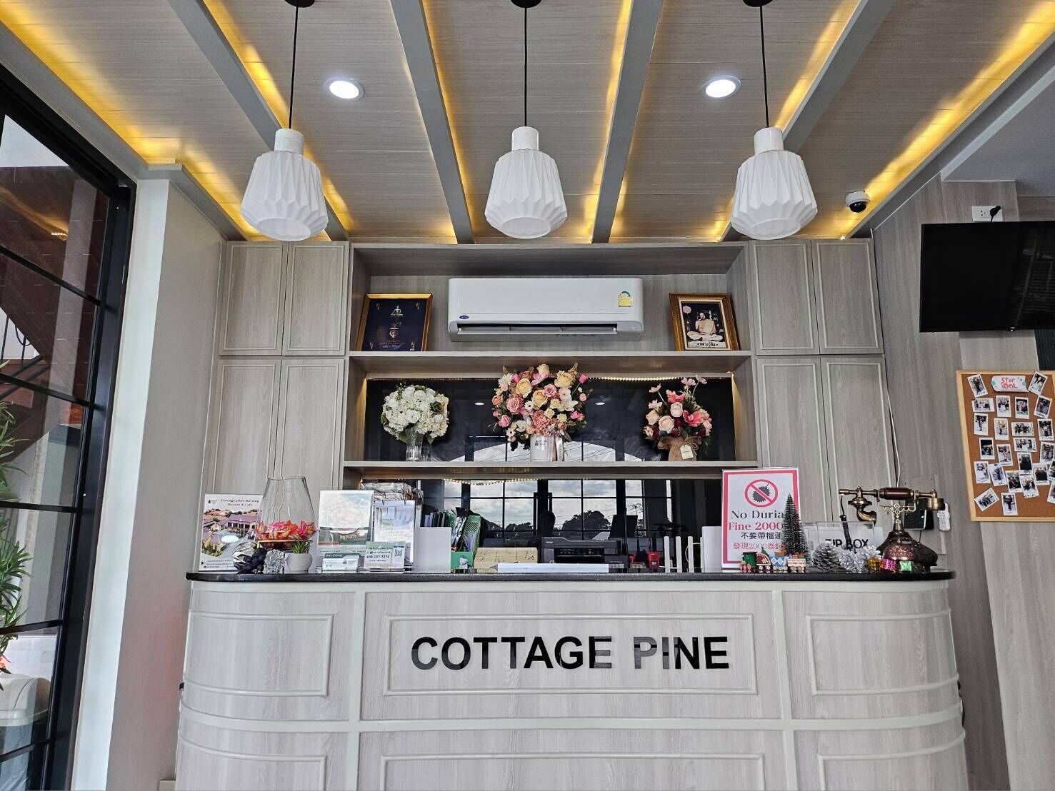 Un comptoir d’accueil portant l’inscription « COTTAGE PINE », agrémenté d’un arrangement floral, d’un appareil de climatisation et d’un panneau indiquant « Interdiction de jeter des objets tranchants ».