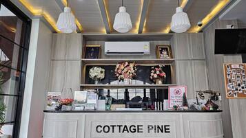 Un comptoir d’accueil portant l’inscription « COTTAGE PINE », agrémenté d’un arrangement floral, d’un appareil de climatisation et d’un panneau indiquant « Interdiction de jeter des objets tranchants ».