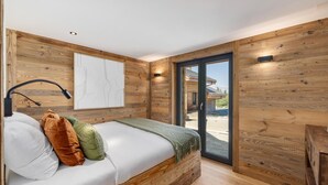 A bedroom with a bed, pillows, a bedside lamp, a wall-mounted artwork, and a view through a glass door to the outside. - Chalet Le Reve Cinema Room Jacuzzi (Riddes)