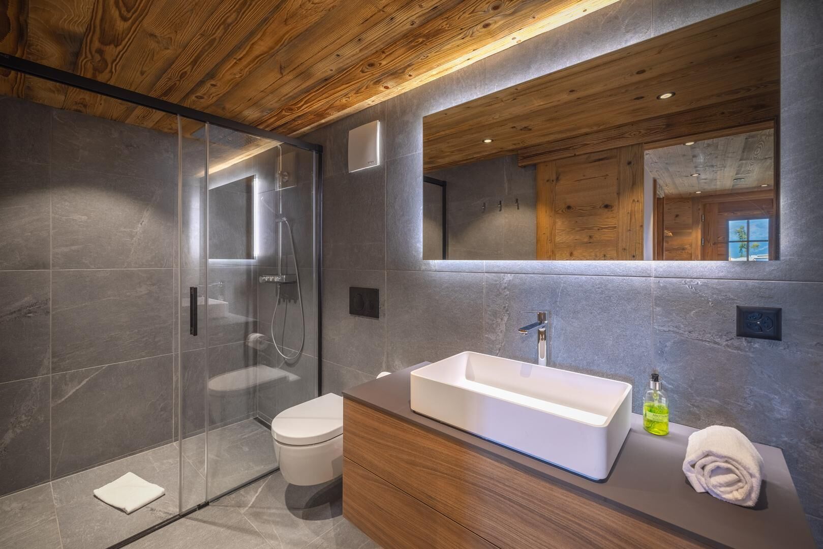 A modern bathroom with a wooden ceiling, a large mirror, a white sink, and a glass-enclosed shower.
