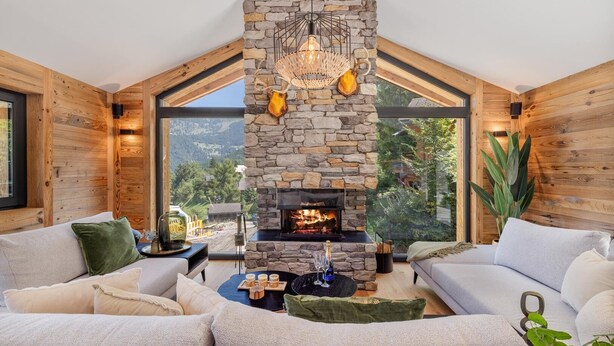 A cozy living room with a stone fireplace, wooden walls, and large windows offering a view of the outdoors. - Chalet Le Reve Cinema Room Jacuzzi (Riddes)