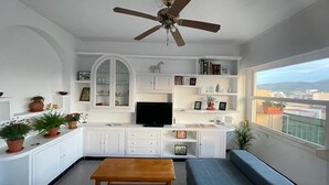 Ein Wohnzimmer mit Fernseher, weißen Schränken, Regalen mit Büchern und Dekorationen, einem Deckenventilator und einem Fenster mit Blick auf die Stadt.