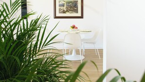 Un coin repas avec une table et des chaises blanches, un tableau encadré au mur et des plantes vertes au premier plan.