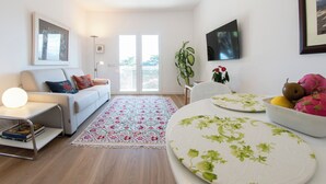 Un salon moderne comprenant un canapé, une table basse, une table à manger avec un panier de fruits, et une grande fenêtre offrant une vue dégagée.