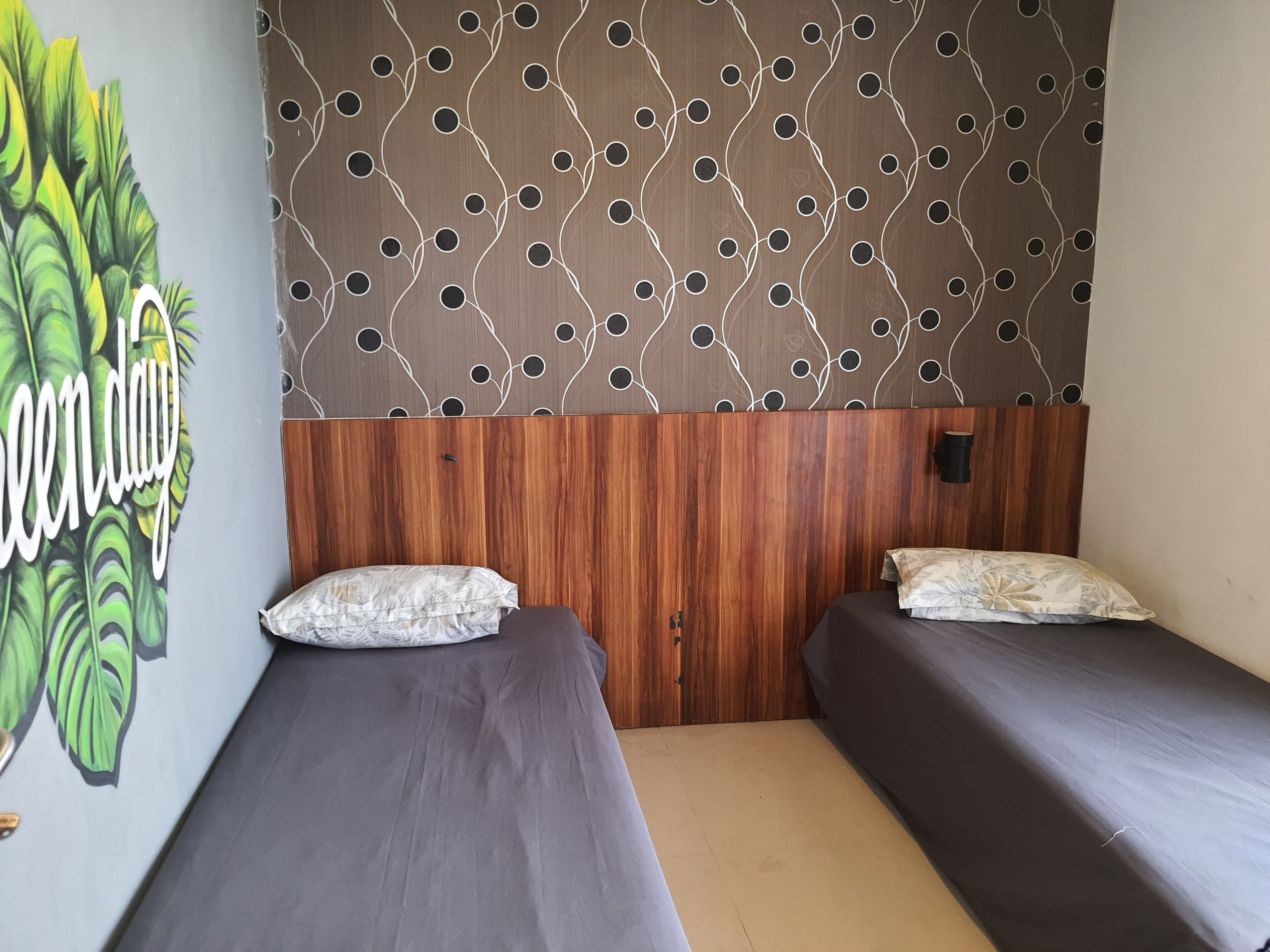 A room with two single beds, a wooden headboard, a wall with a leaf pattern, and a 'green day' mural.