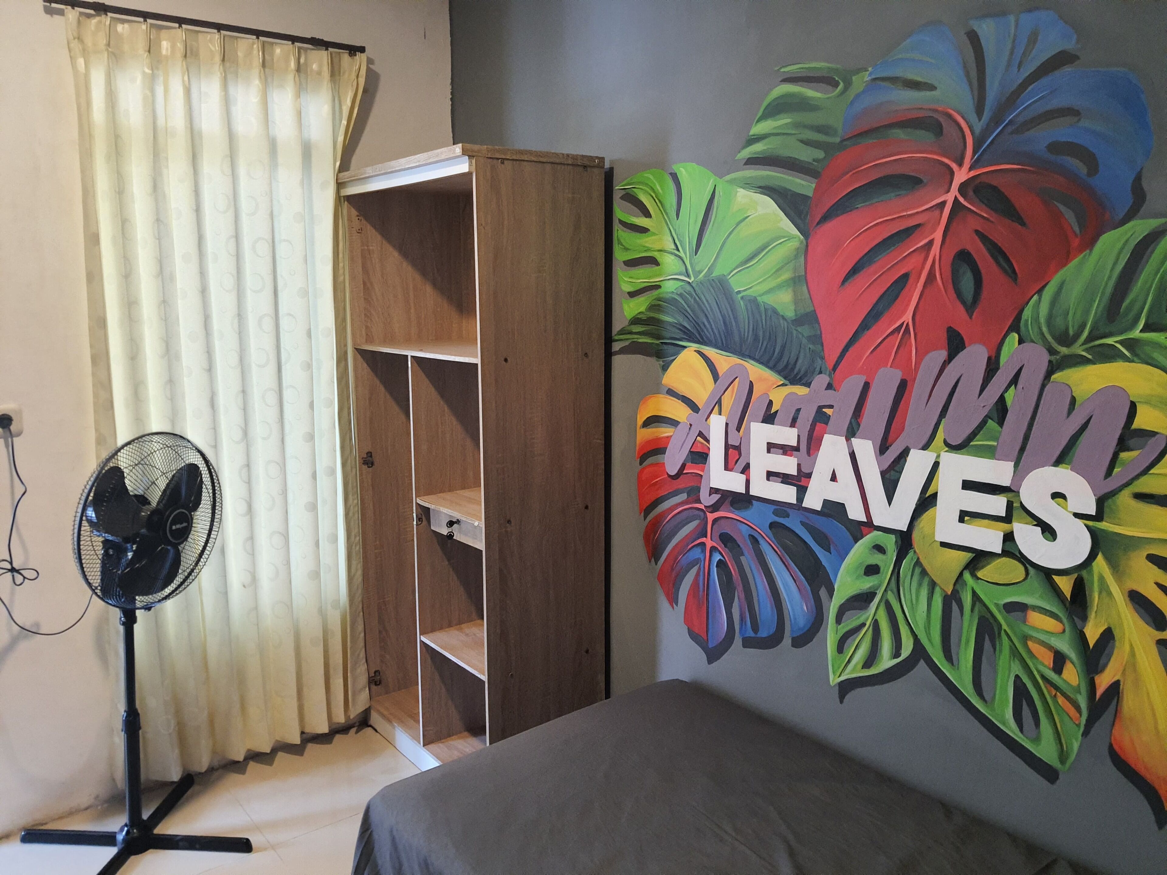 A room with a bed, a wardrobe, a fan, and a wall with a colorful leaf mural.