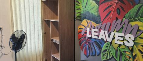 A room with a bed, a wardrobe, a fan, and a wall with a colorful leaf mural.