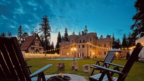 A courtyard with two wooden chairs, a fire pit, and a stone circle. In the background, there is a large stone building with multiple windows and string lights. - The Castle - Your unique way of retreat (Samokov)