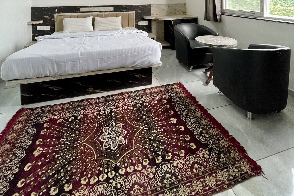 A bedroom with a bed, a ceiling fan, a window with curtains, a small table, a chair, and a decorative rug.
