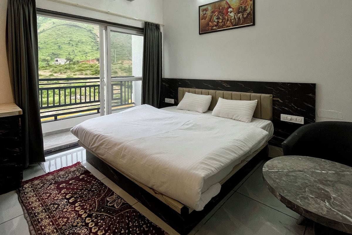 A hotel room with a bed, a ceiling fan, an air conditioning unit, a painting on the wall, a view of greenery outside, and a marble table.