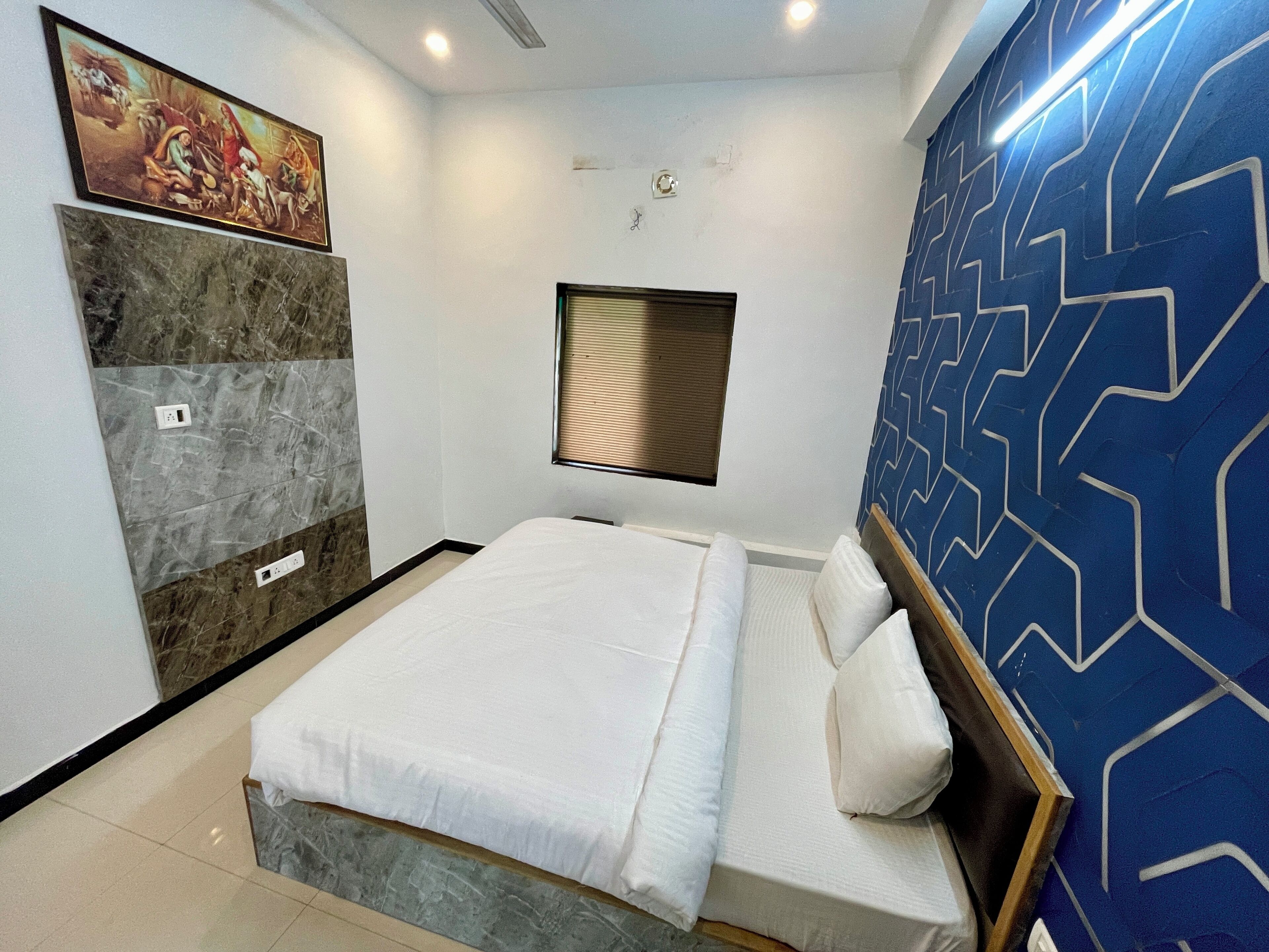 A hotel room with a bed, a wall-mounted painting, a window with blinds, and a blue patterned wall.
