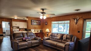 A spacious living room with two leather sofas, a wooden floor, a ceiling fan, and a view of the backyard through a large window. - Skyline Lodge 
Lake Dardanelle 
Arkansas River Valley (Russellville)