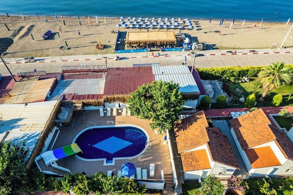 Vista aerea di un resort con piscina, ombrelloni da spiaggia e un edificio con tetto di paglia.