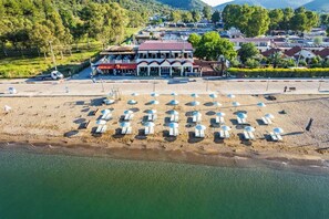 Vista aerea di una spiaggia con file di lettini e ombrelloni, un edificio con tetto rosso e sullo sfondo case residenziali e vegetazione.