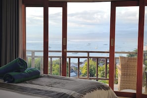 A room with a large window overlooking the sea, a bed with a striped blanket, and a wicker chair.