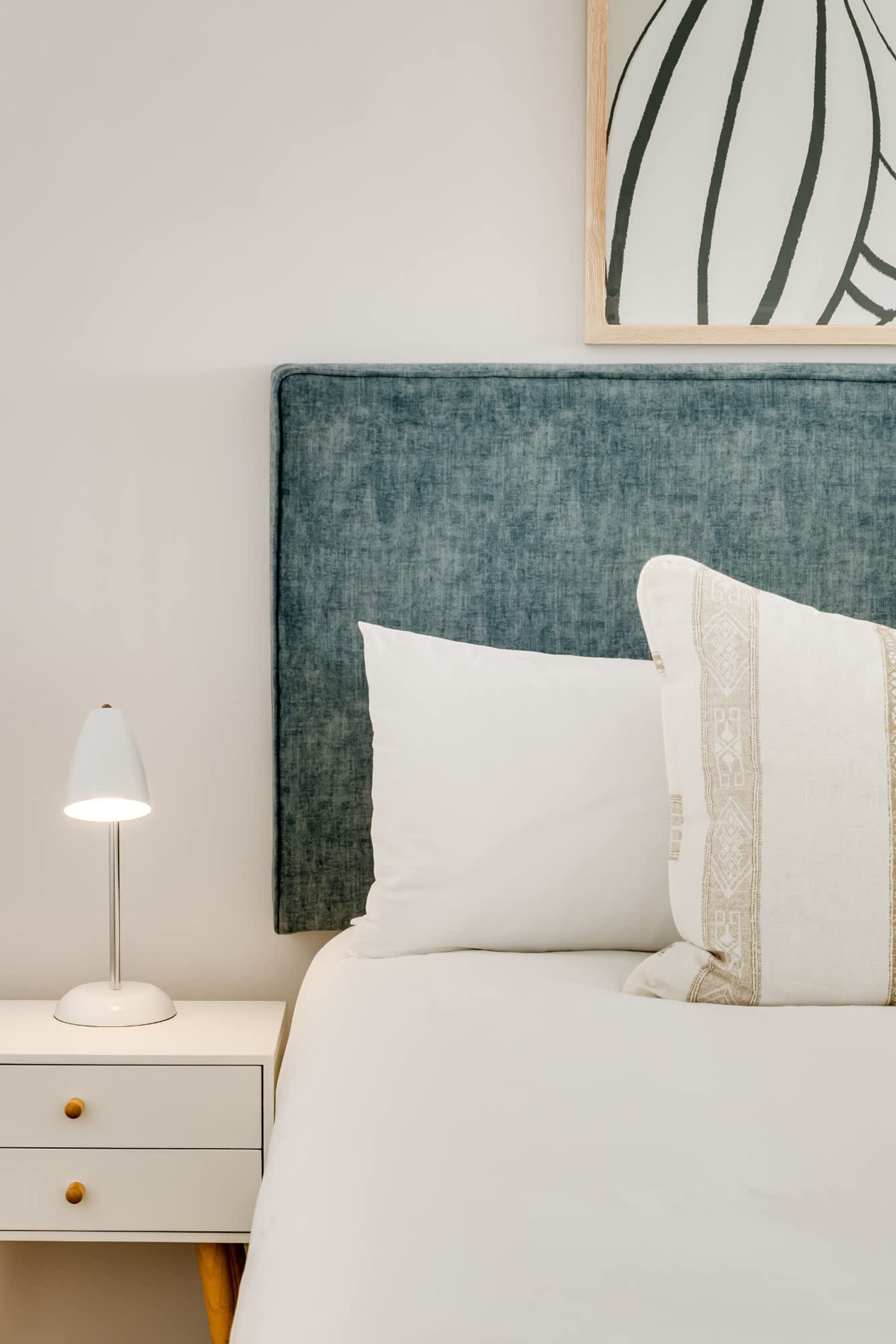 A neatly made bed with white bedding and decorative pillows, a bedside lamp, and a wall-mounted artwork.