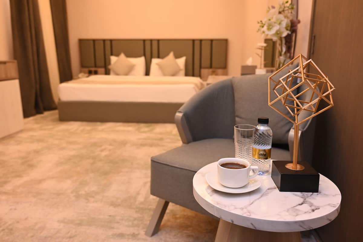 Una camera d'albergo con un letto, una poltrona grigia, un tavolino rotondo con una tazza di caffè, una bottiglia d'acqua e un oggetto decorativo geometrico.