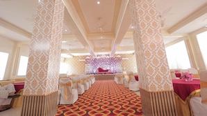 Une salle de banquet dotée d’une scène, de rangées de tables recouvertes de nappes blanches et ornées de rubans dorés, ainsi que de colonnes à motifs.