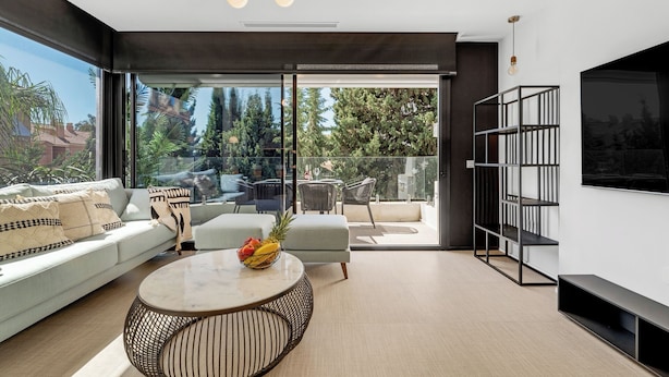 A modern living room with a sofa, a round coffee table with fruit, a dining area with chairs, a bookshelf, and a TV mounted on the wall. - Dunes Paradise Suite (Marbella)