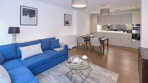 A modern living room with a blue sofa, a glass coffee table, and a dining area with chairs and a table. - Modern, Luxurious 1BR Flat- Heart of Covent Garden (London)