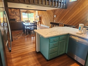 Una cocina con encimera de mármol, armarios color turquesa y lavavajillas. Hay una zona de comedor con una mesa y sillas, y una chimenea de piedra al fondo.