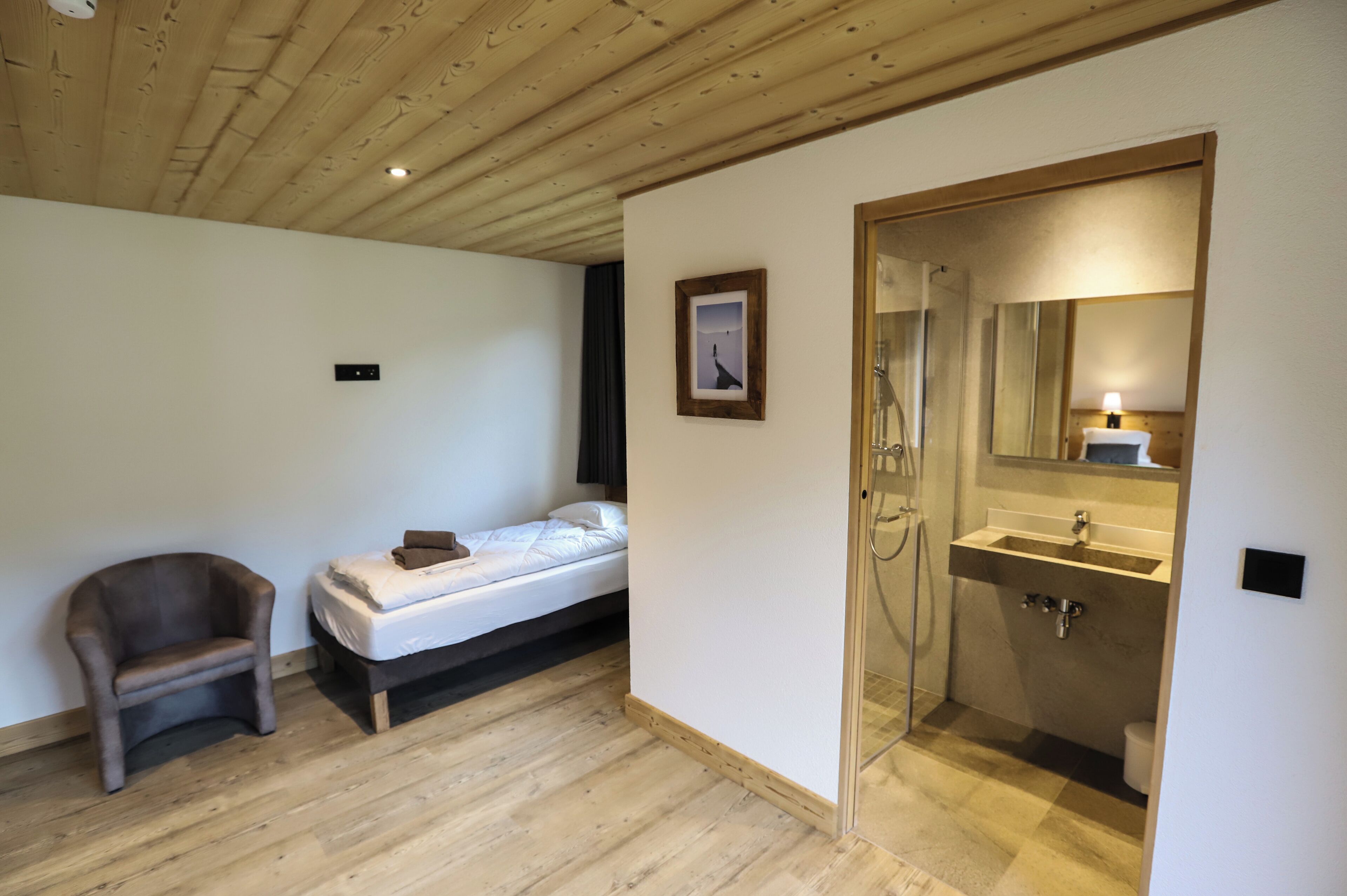 A bedroom with a bed, a chair, a bedside table, a framed picture, and a bathroom with a sink and mirror.