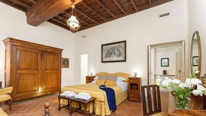 A bedroom with a large wooden wardrobe, a bed with a yellow bedspread, a bedside table with a lamp, a chair, and a vase with flowers. - Cartari Deluxe Apartment (Rome)