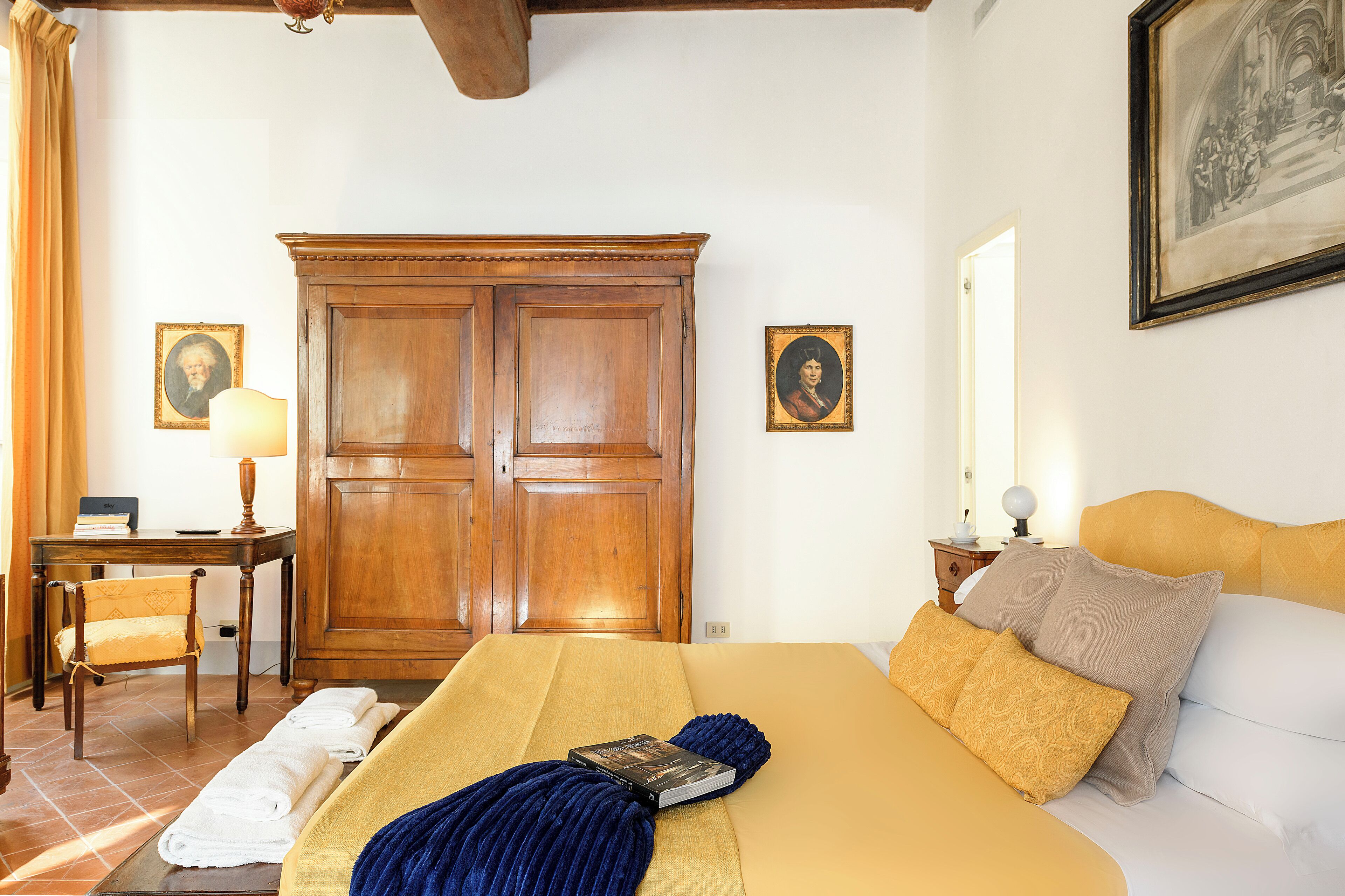 Una habitación de hotel con una cama, un armario de madera, un escritorio con lámpara y dos retratos enmarcados en la pared.