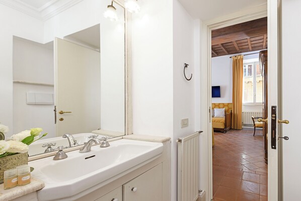 A bathroom with a white sink, mirror, and a door leading to a room with a bed and a window. - Cartari Deluxe Apartment (Rome)