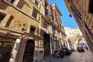 A street view with a barber shop on the left, a building with a balcony, and parked cars. - Cartari Deluxe Apartment (Rome)