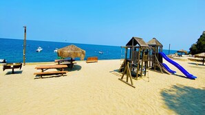A beach with a wooden play structure, a blue slide, picnic tables, and a thatched umbrella.