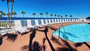 A poolside area with white lounge chairs and palm trees, overlooking a clear blue ocean.
