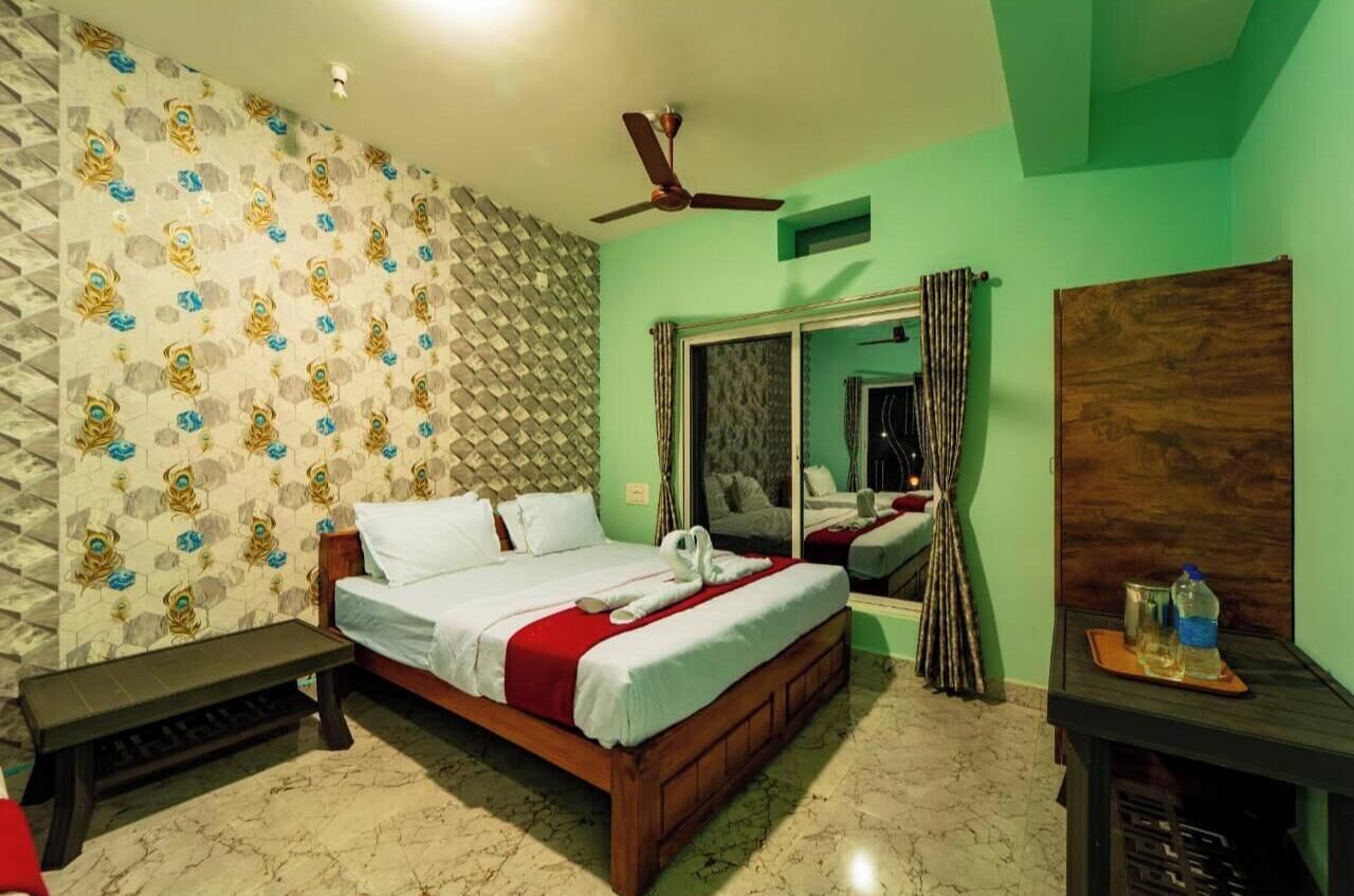 A hotel room with a bed, a ceiling fan, a mirror, and a wooden table with a bottle and glasses.