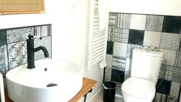 A bathroom with a round white sink, black faucet, and a wooden countertop. There is a toilet with a white tank and a black trash can beside it. The floor has hexagonal tiles, and the walls have a black and white patterned backsplash.