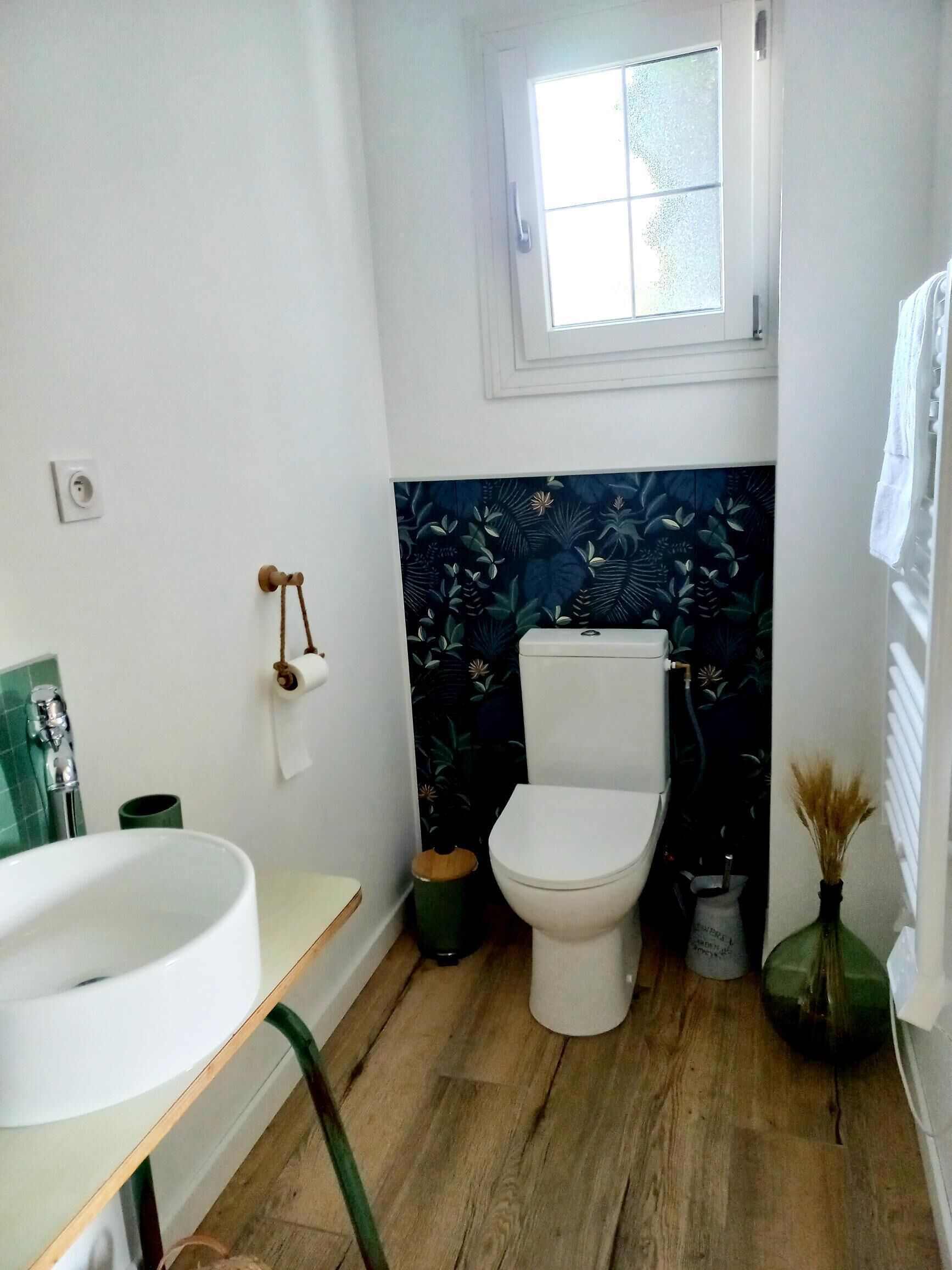 Ein Badezimmer mit Toilette, grünem Mülleimer, Waschbecken und einem Fenster über der Toilette.