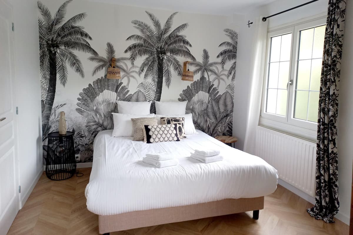 A bedroom with a large bed, a wall mural of palm trees, a window with curtains, and a small bedside table with a lamp.