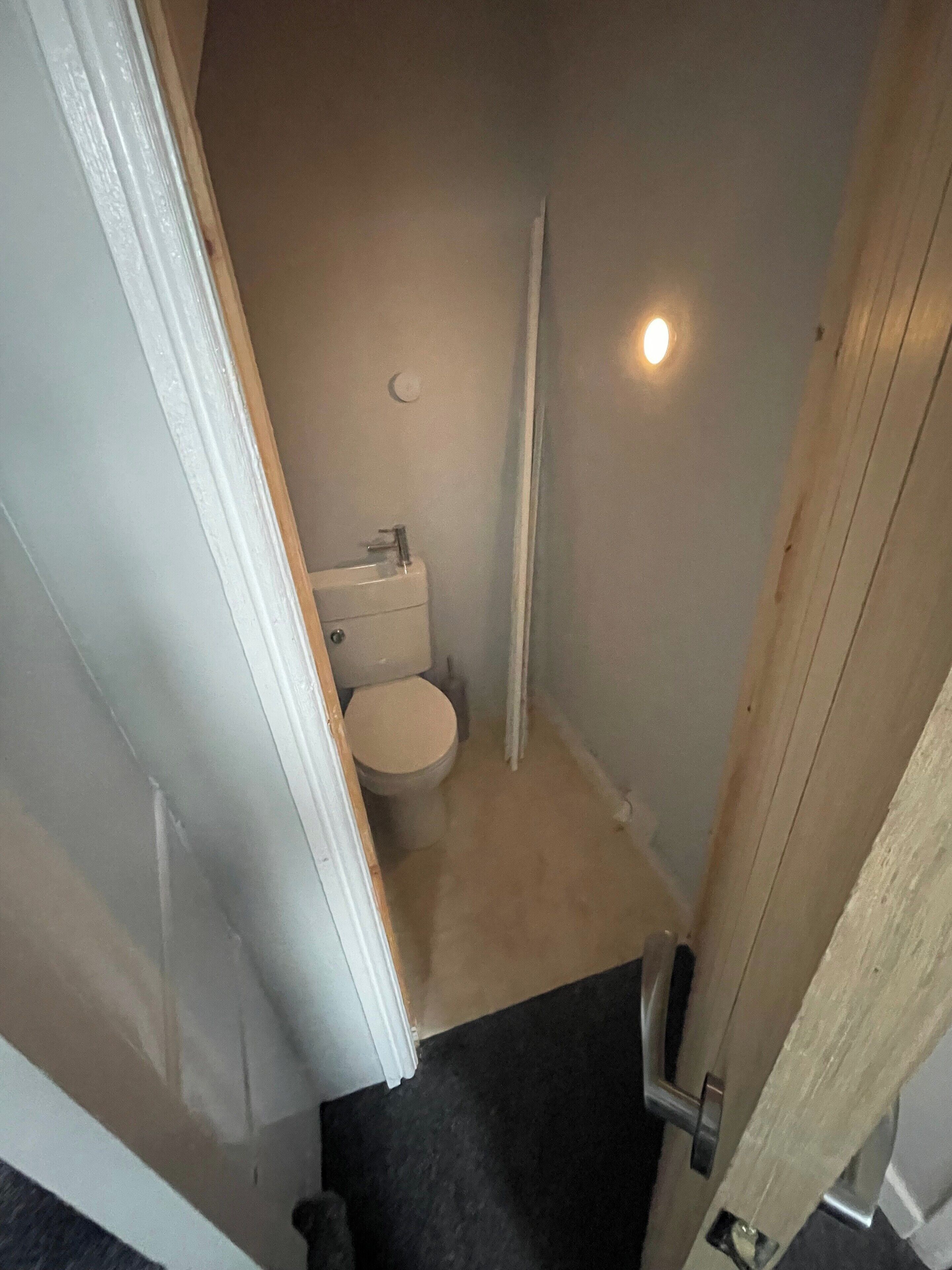 A narrow bathroom with a toilet, a small sink, and a door leading to another room.